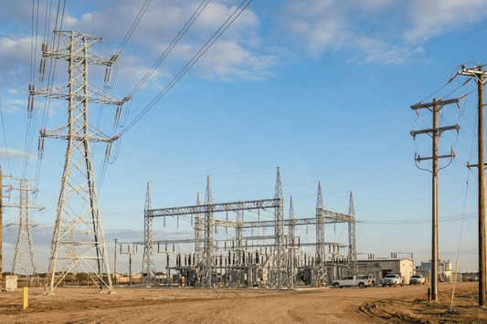 Power substation with steel transmission towers and wooden utility poles under a blue sky, dirt road and service vehicles nearby