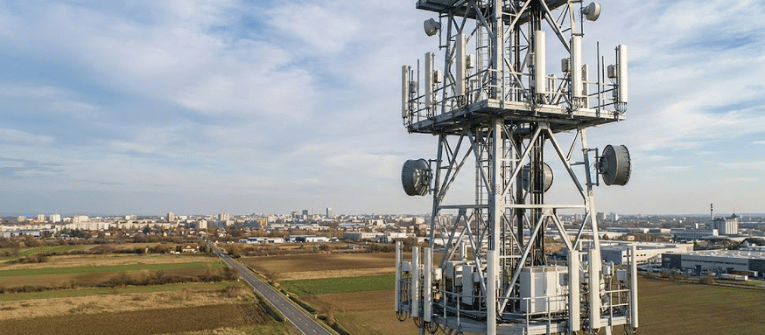 Telecom tower with antennas overlooking farmland and a distant city skyline under a partly cloudy sky, road leading toward city